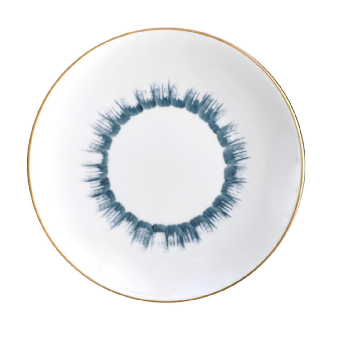 The Marie Daâge Iris 1 Dinnerware by Marie Daâge features a round white Limoges porcelain plate with a thin gold rim and a blue brushstroke-style circular pattern at the center, evoking a ring or sunburst.
