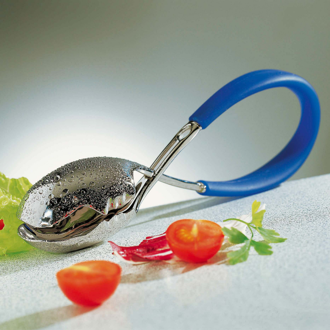 The Casa Bugatti KISS Salad Tong, with its shiny metal finish and blue handle, rests on a light surface beside cherry tomatoes, red chili pepper pieces, parsley, and lettuce—a stylish accessory for summer entertaining.