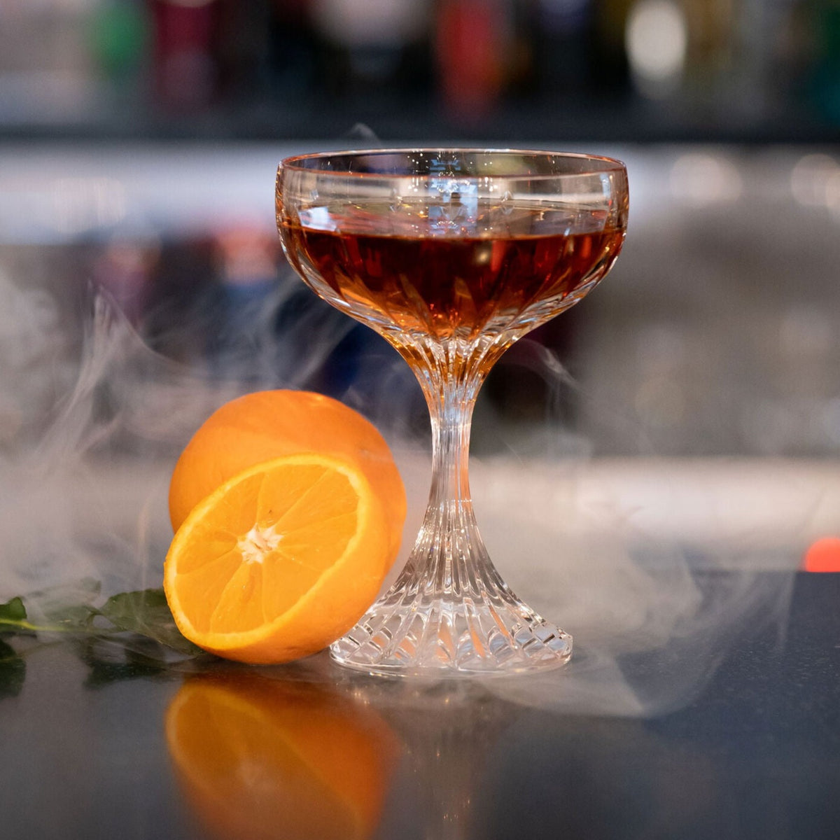The Masséna Coupe Set of 2 by Baccarat features elegant glasses filled with a dark amber cocktail beside a halved orange and wisps of smoke—making it a striking upgrade to any barware collection.