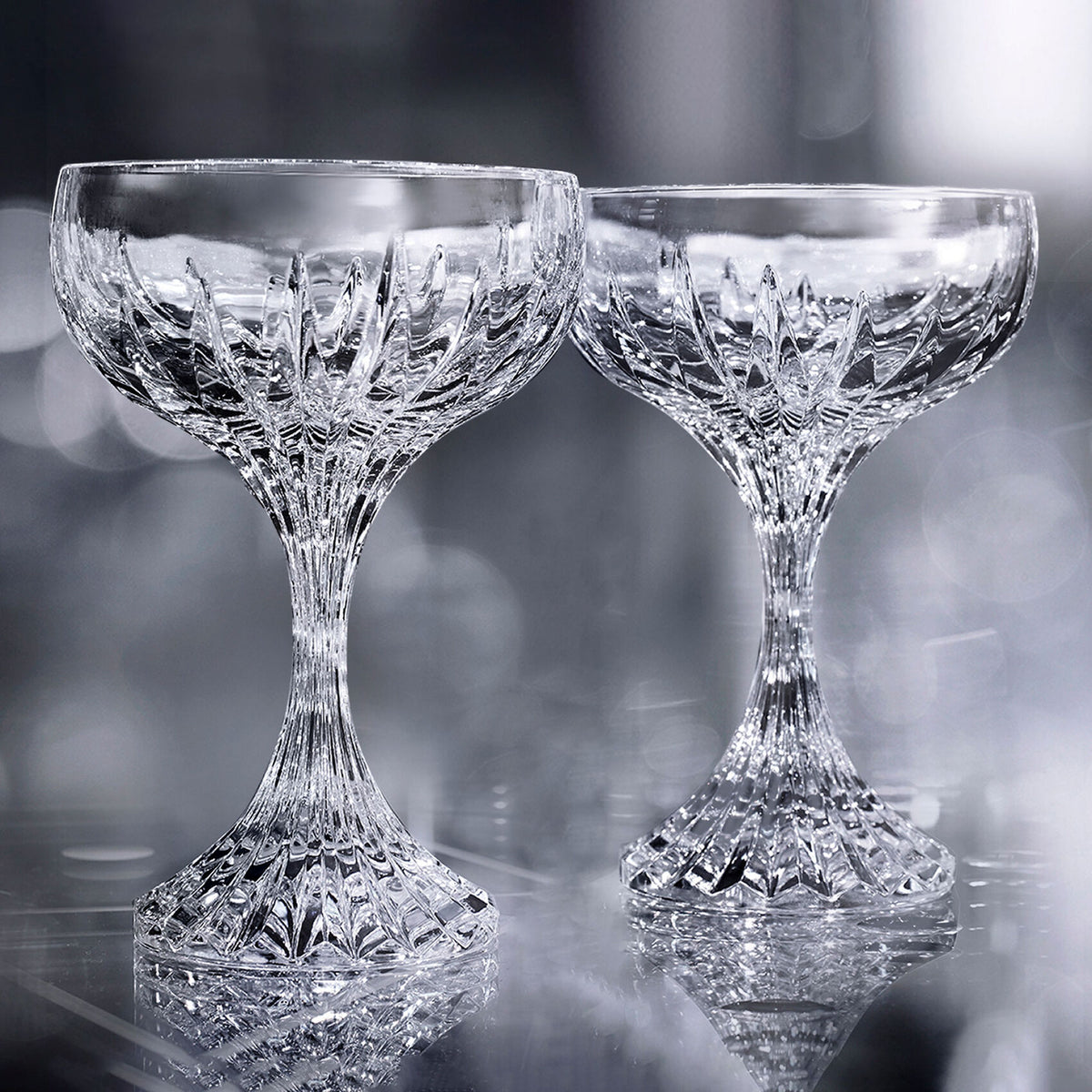 Two Masséna Coupe glasses from Baccarat stand side by side on a reflective surface, their intricate crystal patterns highlighting the elegance of this refined barware set of 2 against a soft, silvery backdrop.