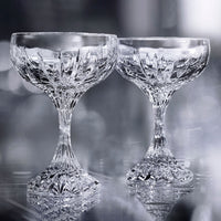 Two Masséna Coupe glasses from Baccarat stand side by side on a reflective surface, their intricate crystal patterns highlighting the elegance of this refined barware set of 2 against a soft, silvery backdrop.