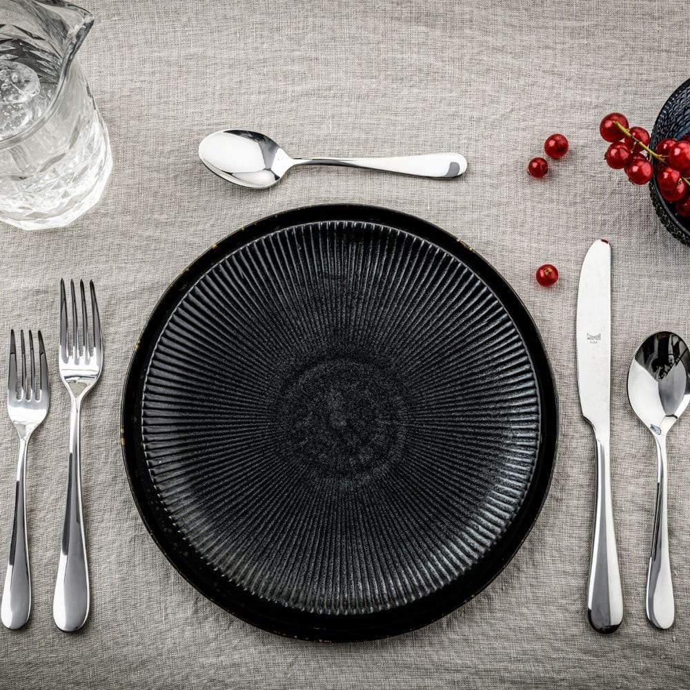 A black textured plate rests on a beige tablecloth, surrounded by Mepra's Natura Flatware 5 Piece Setting in stainless steel, a glass pitcher, and a cluster of red berries arranged in the top right corner.
