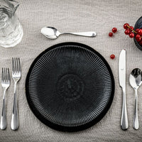 A black textured plate rests on a beige tablecloth, surrounded by Mepra's Natura Flatware 5 Piece Setting in stainless steel, a glass pitcher, and a cluster of red berries arranged in the top right corner.