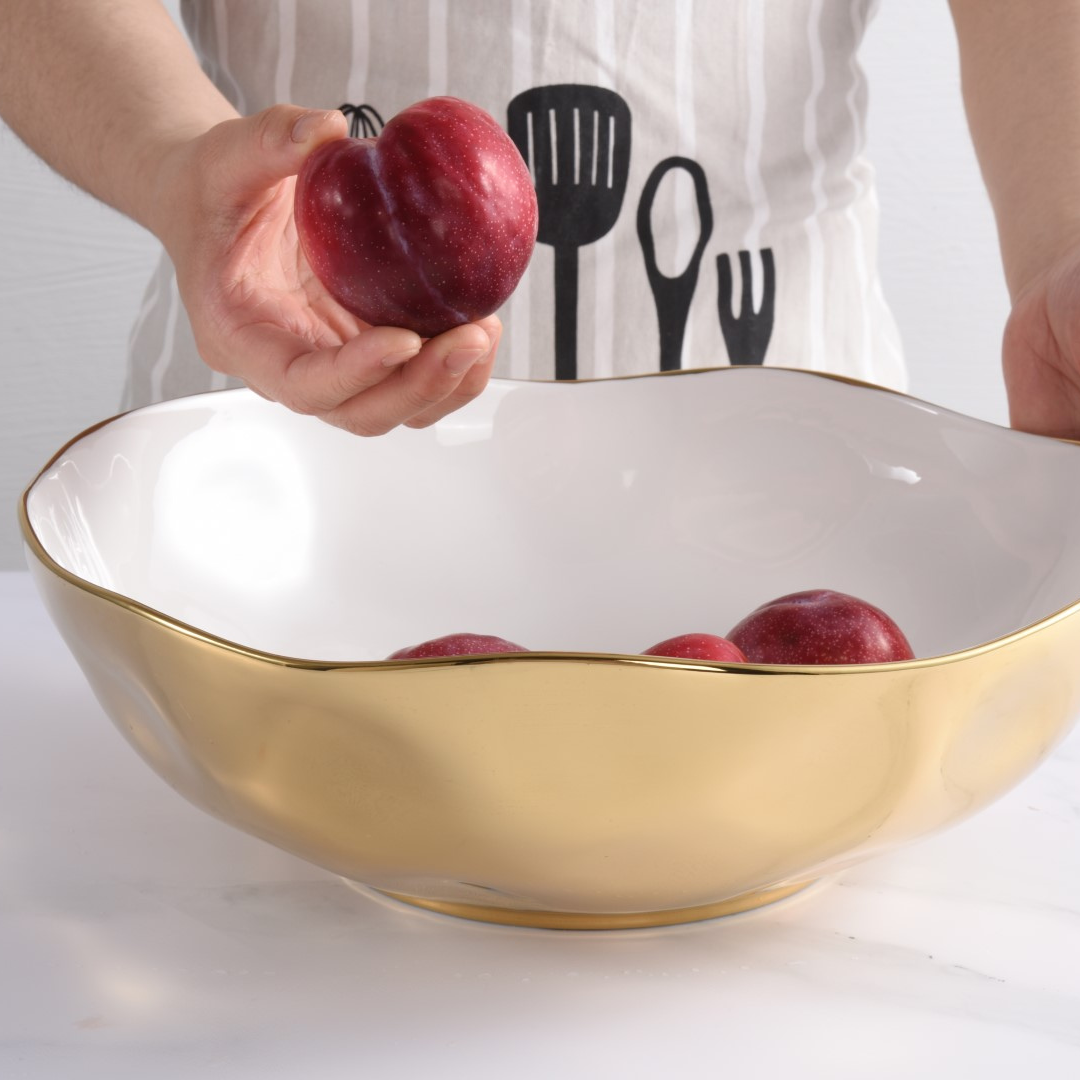 A person in a utensil-print apron holds a red plum above the PAMPA BAY Moonlight Bowl, a large white porcelain serveware bowl with a gold titanium rim, filled with more plums.