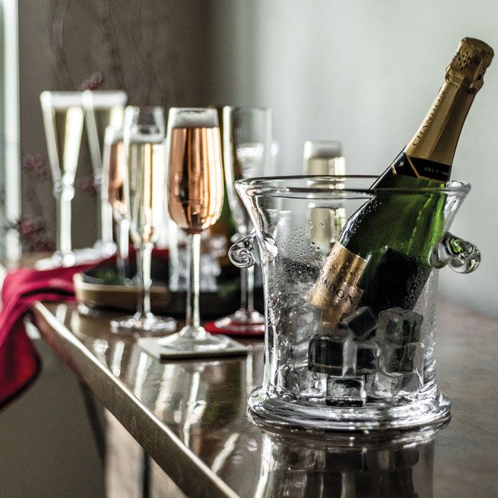 A Norwich Ice Bucket by Simon Pearce, filled with ice and a champagne bottle, sits on a table surrounded by filled champagne flutes and wine glasses, with a red cloth in the background.