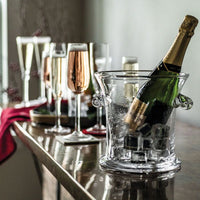 A Norwich Ice Bucket by Simon Pearce, filled with ice and a champagne bottle, sits on a table surrounded by filled champagne flutes and wine glasses, with a red cloth in the background.