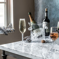 A marble table displays a Simon Pearce Norwich Ice Bucket chilling champagne, alongside a flute, two wine glasses, and a wine bottle. Pinecones and frosted pine branches decorate the setting as light pours in from a nearby window.