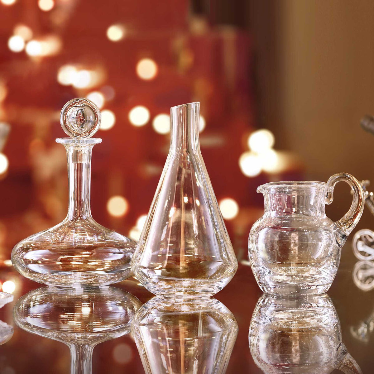 The Baccarat Oenologie Decanter by Baccarat is showcased on a reflective surface, accompanied by a carafe and small pitcher, with warm, blurred festive lights in the background for an elegant effect.