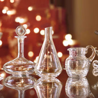 The Baccarat Oenologie Decanter by Baccarat is showcased on a reflective surface, accompanied by a carafe and small pitcher, with warm, blurred festive lights in the background for an elegant effect.