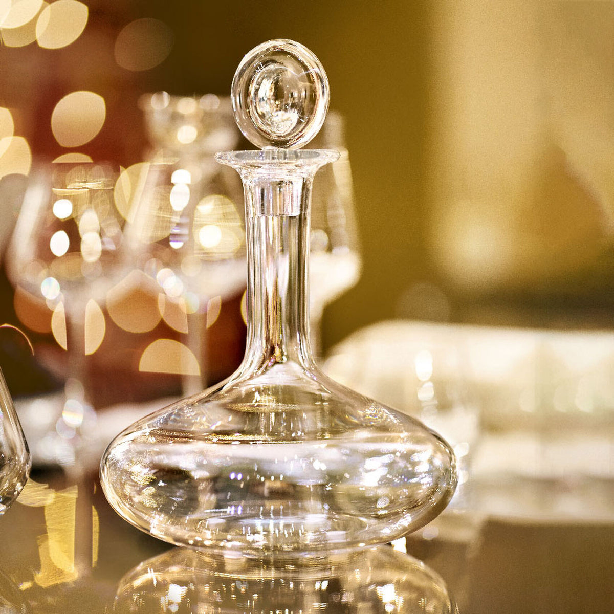 The Baccarat Oenologie Decanter by Baccarat, featuring a round stopper, stands on a reflective surface with wine glasses and warm bokeh lights blurred in the background.