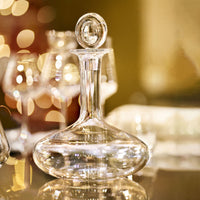 The Baccarat Oenologie Decanter by Baccarat, featuring a round stopper, stands on a reflective surface with wine glasses and warm bokeh lights blurred in the background.
