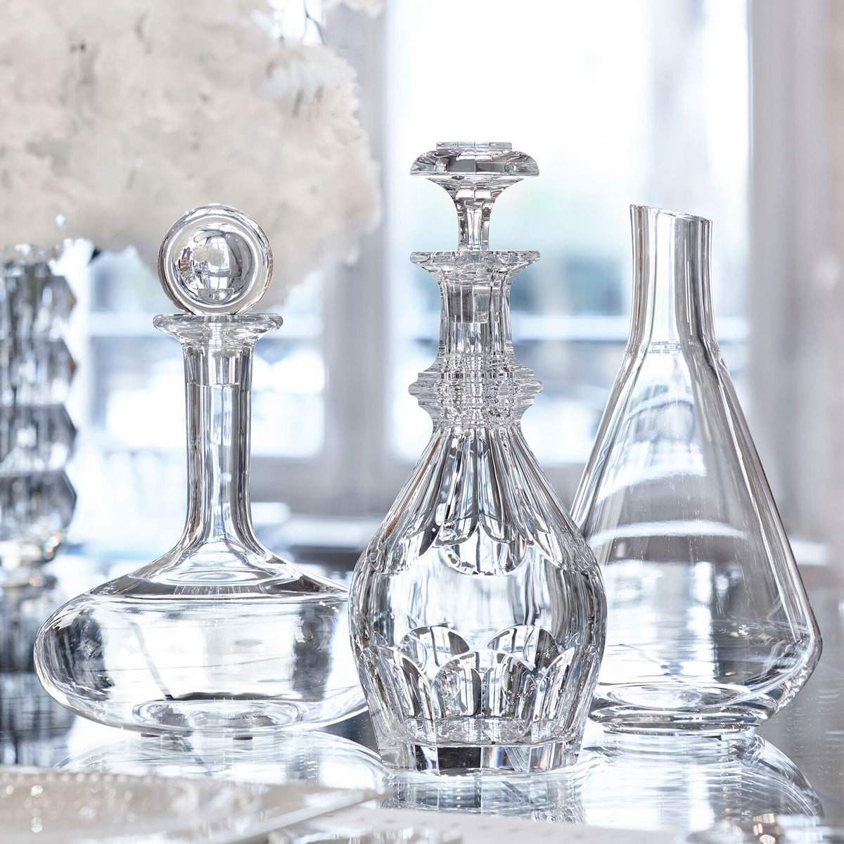 The Baccarat Oenologie Decanter by Baccarat, featuring exquisite craftsmanship, is showcased on a reflective surface with blurred floral decor and large bright windows in the background.