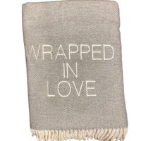 The Wrapped in Love Throw by A Soft Idea is a folded light gray blanket with white fringe and "WRAPPED IN LOVE" in bold white letters—super soft, machine washable, and a perfect holiday gift.