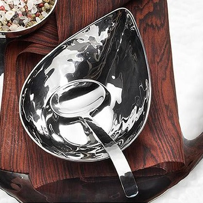 The Mary Jurek Valencia Gravy Ladle sits in a curved, handmade stainless steel serving bowl on a folded brown napkin atop dark wood. A bowl of coarse salt or spices is partially seen in the corner.