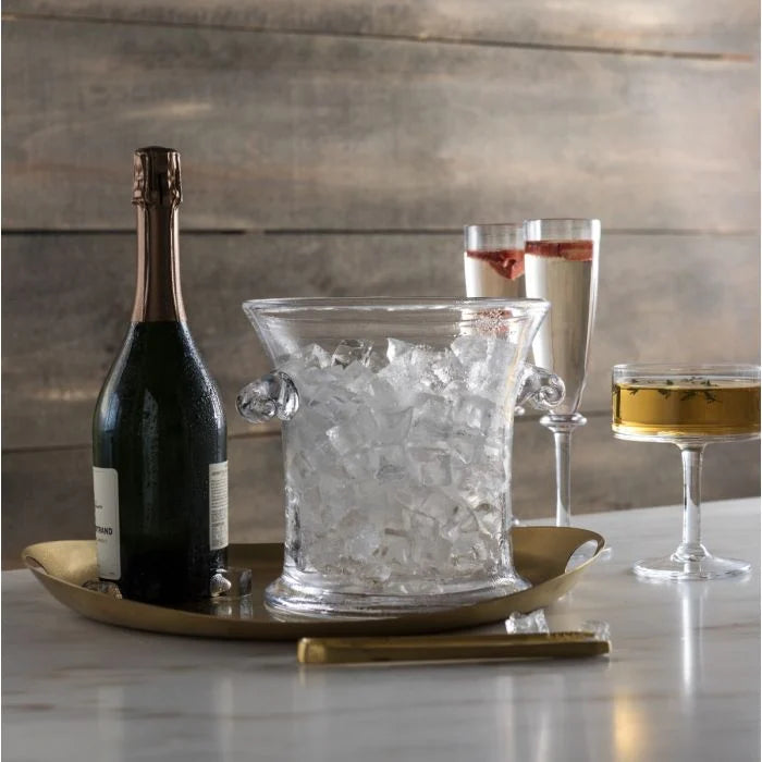A bottle of champagne chills in the Simon Pearce Norwich Ice Bucket filled with ice, accompanied by two drinks on a round brass tray atop a marble surface, set against a wooden background.
