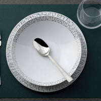A Rosenthal Skin Platinum TAC 02 dinnerware bowl with a geometric rim holds a silver spoon and sits on a matching plate; a glass plus fork and knife appear on a textured green placemat. Made in Germany.