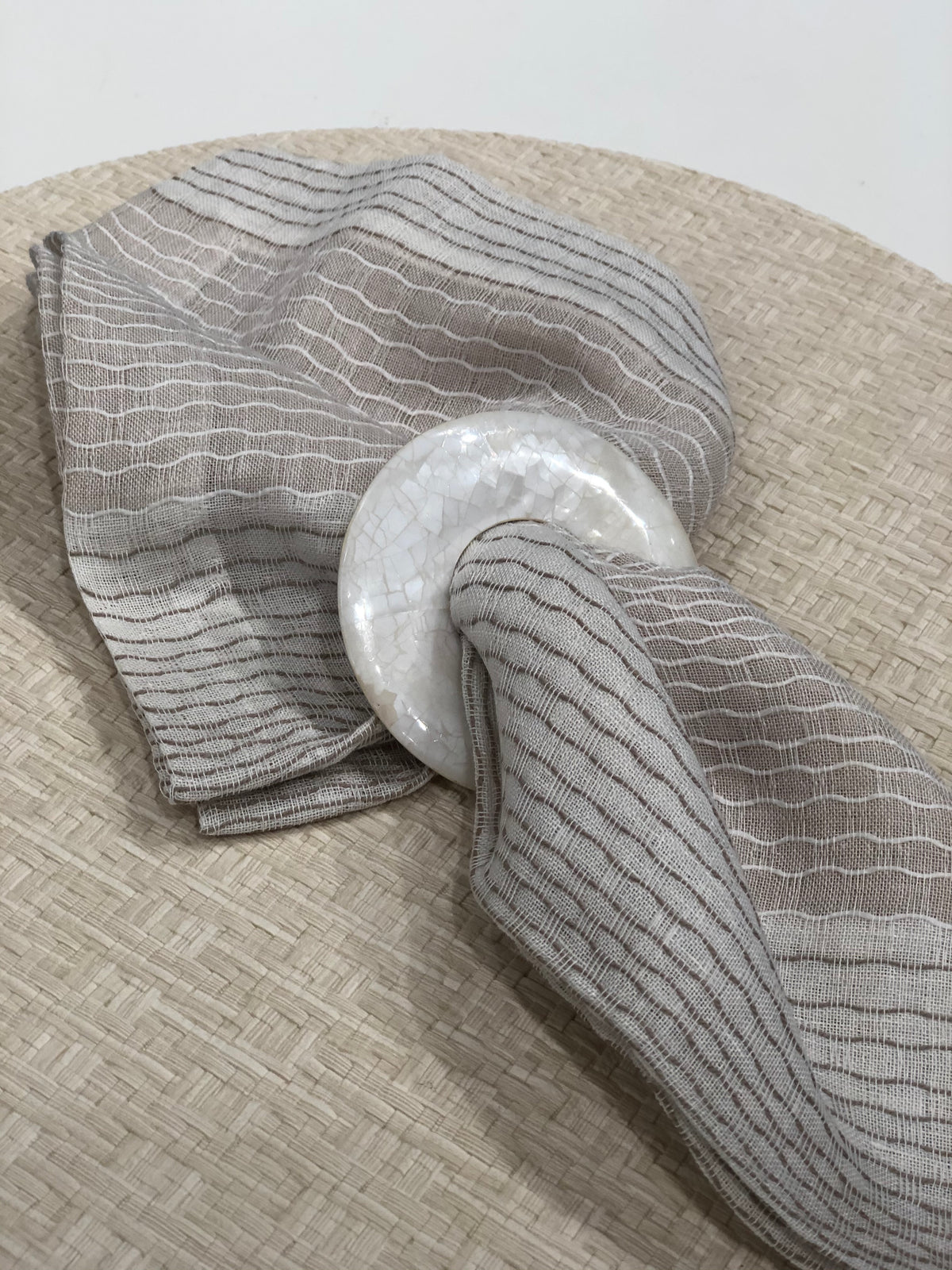A beige and gray striped cloth napkin with a white, round, textured ring rests on Bodrum's Wicker Round Placemat Cream (Set of 4)—a chic accent for outdoor dining.