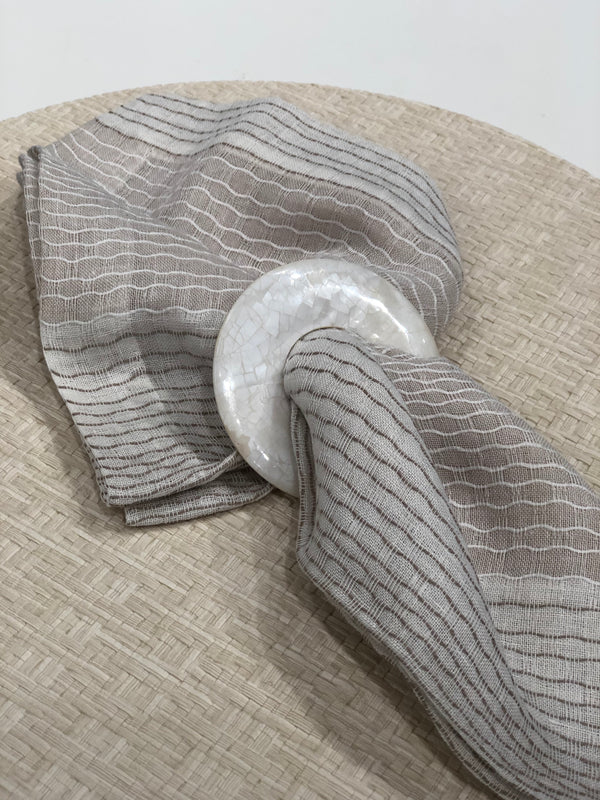 A beige and gray striped cloth napkin with a white, round, textured ring rests on Bodrum's Wicker Round Placemat Cream (Set of 4)—a chic accent for outdoor dining.