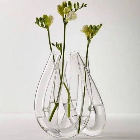 Three green-stemmed flowers are displayed in the Trui Vase by Rosenthal, featuring three connected, looped chambers partially filled with water, set against a plain white background.
