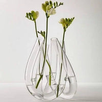 Three green-stemmed flowers are displayed in the Trui Vase by Rosenthal, featuring three connected, looped chambers partially filled with water, set against a plain white background.