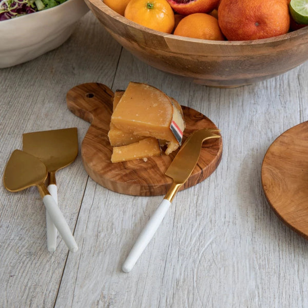 A wooden turtle-shaped cutting board holds a wedge of hard cheese on a light wood table, next to Be Home's White & Gold Cheese Set and a large bowl of assorted fruit.
