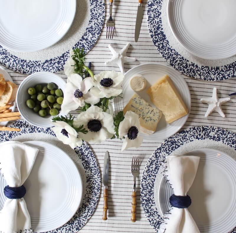A table set for four with white plates on the Splatter Beaded Placemat Set of 4 by Von Gern Home, white cloth napkins with blue rings, cheeses, green olives, white flowers, and starfish atop a striped tablecloth.