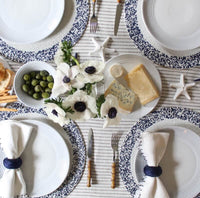 A table set for four with white plates on the Splatter Beaded Placemat Set of 4 by Von Gern Home, white cloth napkins with blue rings, cheeses, green olives, white flowers, and starfish atop a striped tablecloth.