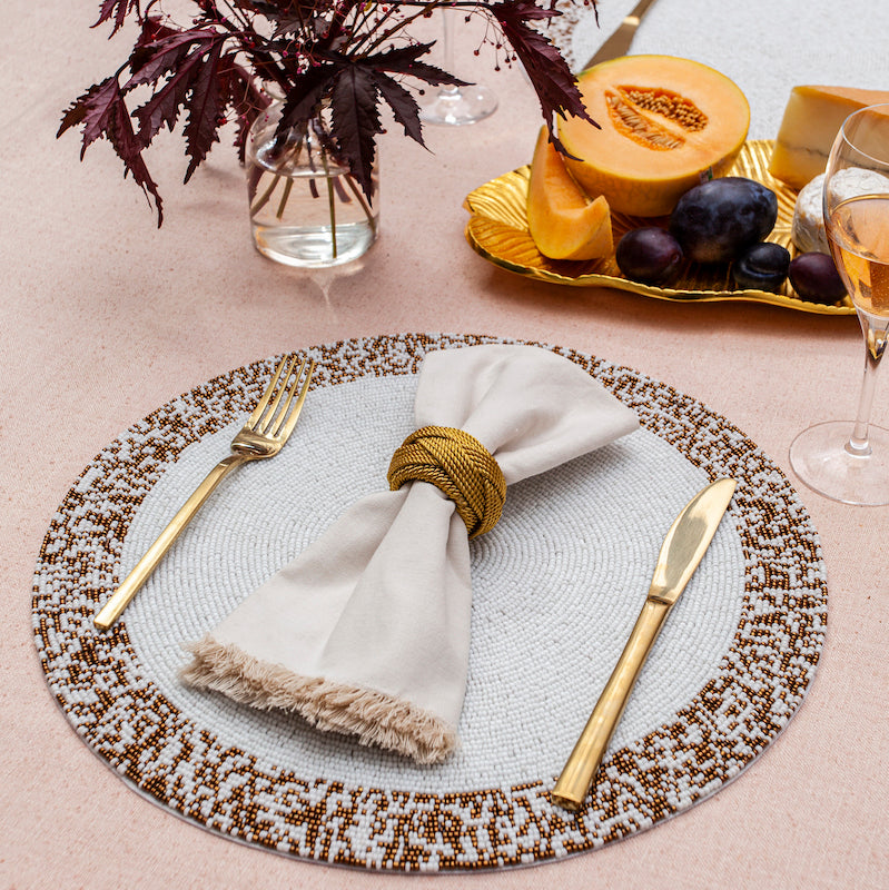The Splatter Beaded Placemat Set of 4 by Von Gern Home displays a gold fork, knife, and fringed beige napkin in a gold ring on a pink tablecloth, with glasses, dark-leaf vase, and melon and grapes nearby.