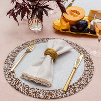 The Splatter Beaded Placemat Set of 4 by Von Gern Home displays a gold fork, knife, and fringed beige napkin in a gold ring on a pink tablecloth, with glasses, dark-leaf vase, and melon and grapes nearby.