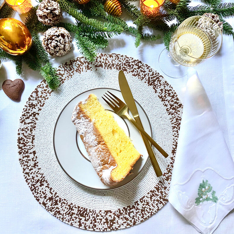 A festive table is set with a slice of sponge cake, gold cutlery, an embroidered napkin, white wine, evergreens, pine cones, ornaments, candles, and Splatter Beaded Placemat Set of 4 by Von Gern Home for added charm.