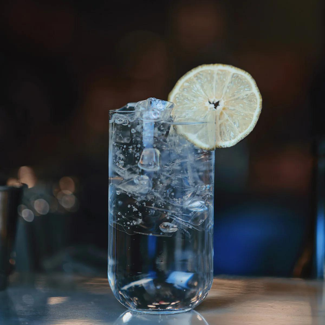 A Luigi Bormioli Sublime Beverage Glass from the set of 4, filled with sparkling water, ice cubes, and garnished with a lemon slice on the rim, rests on a reflective surface against a dark, blurred background.