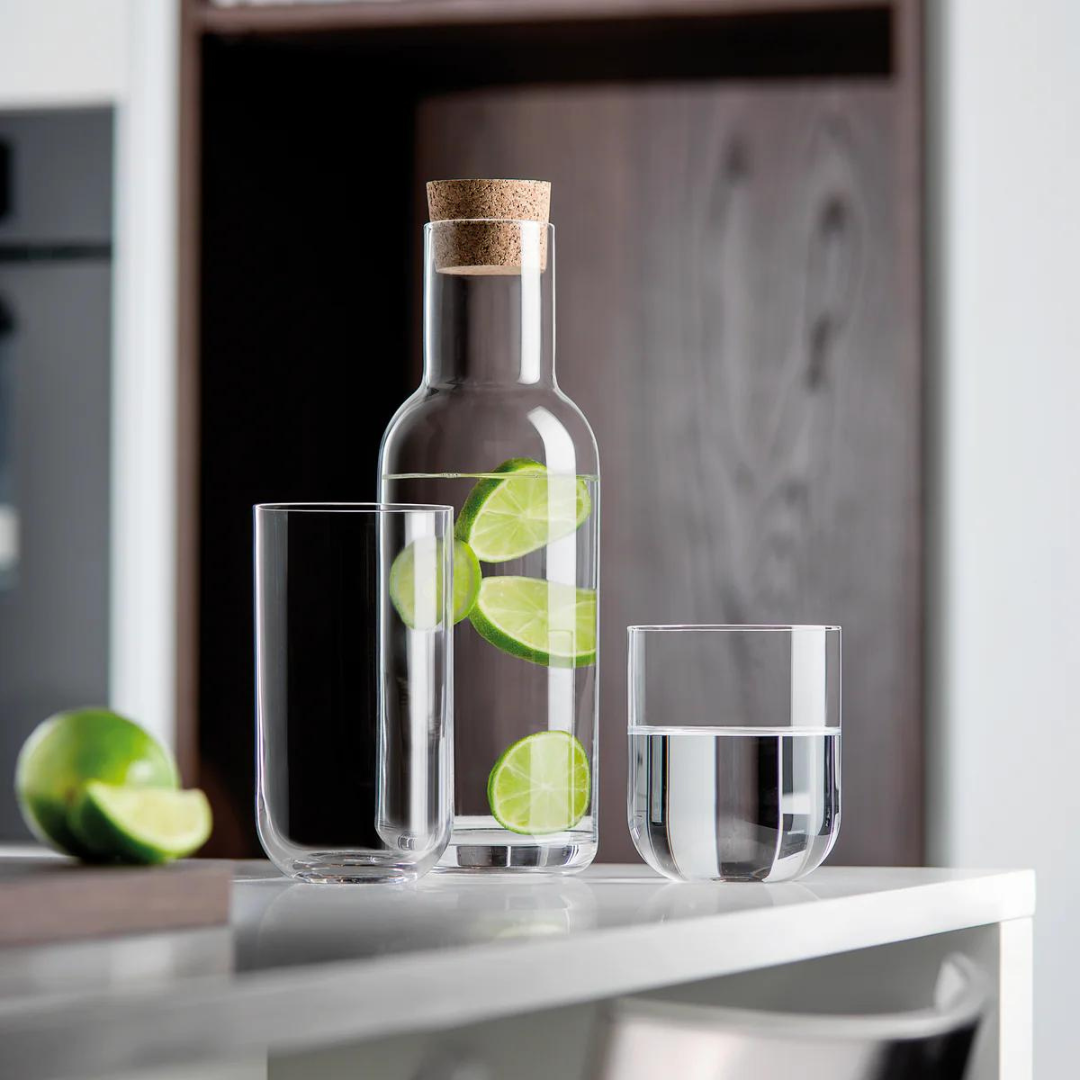 A Luigi Bormioli carafe with cork lid, filled with water and lime slices, stands beside two Luigi Bormioli Sublime Beverage Glasses—one empty, one half-full from the set of 4. A sliced lime sits in the background.