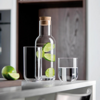 A Luigi Bormioli carafe with cork lid, filled with water and lime slices, stands beside two Luigi Bormioli Sublime Beverage Glasses—one empty, one half-full from the set of 4. A sliced lime sits in the background.