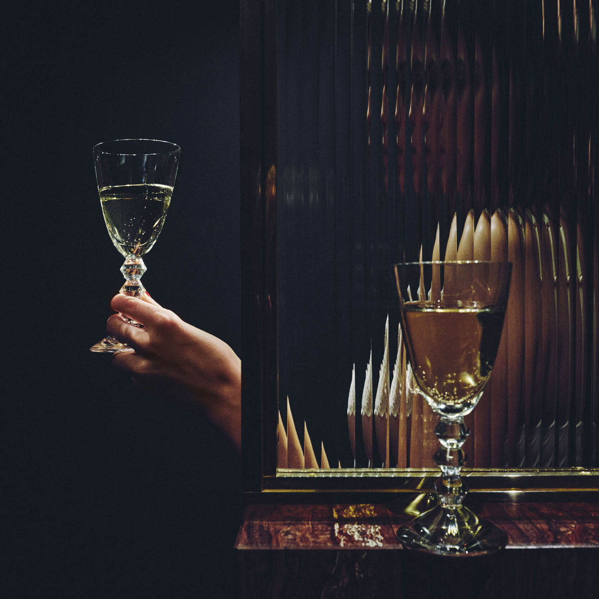 A hand holds the Baccarat Vega White Wine Glass next to a textured glass panel. Behind the panel, a distorted reflection reveals a person and another wine glass on a wooden surface against a dark background.