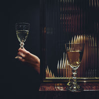 A hand holds the Baccarat Vega White Wine Glass next to a textured glass panel. Behind the panel, a distorted reflection reveals a person and another wine glass on a wooden surface against a dark background.