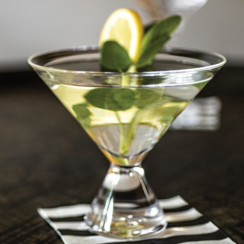 A Simon Pearce Westport Stemless Martini glass holds a clear yellow-green cocktail garnished with lemon and mint, resting on a black and white striped napkin.